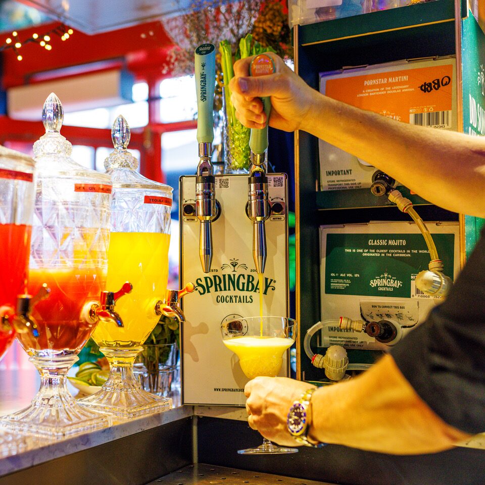 Bartender pours a yellow cocktail from a Springbay tap beside the bag-in-box displays